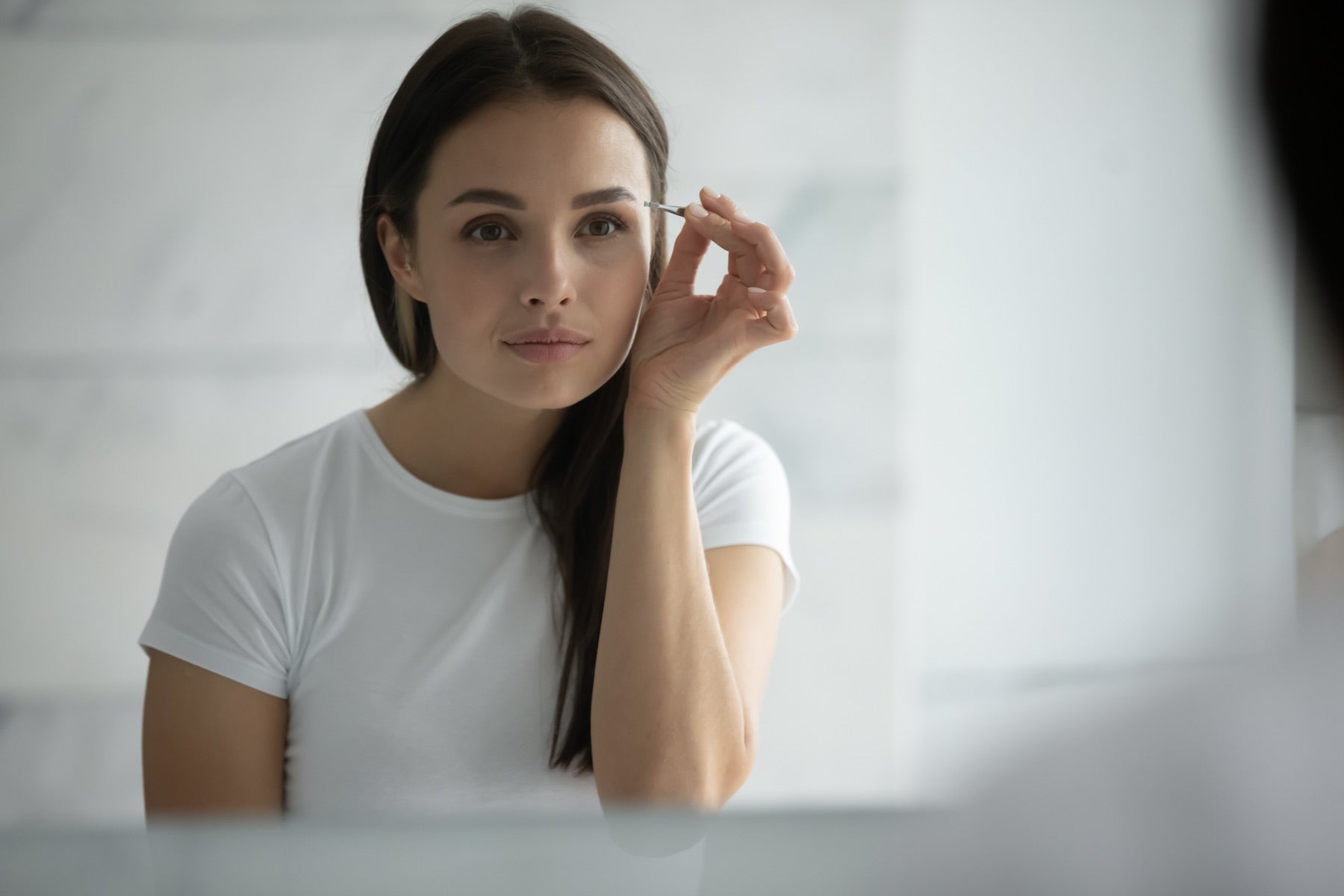 brunette white woman looking in a mirror while tweezing her eyebrows